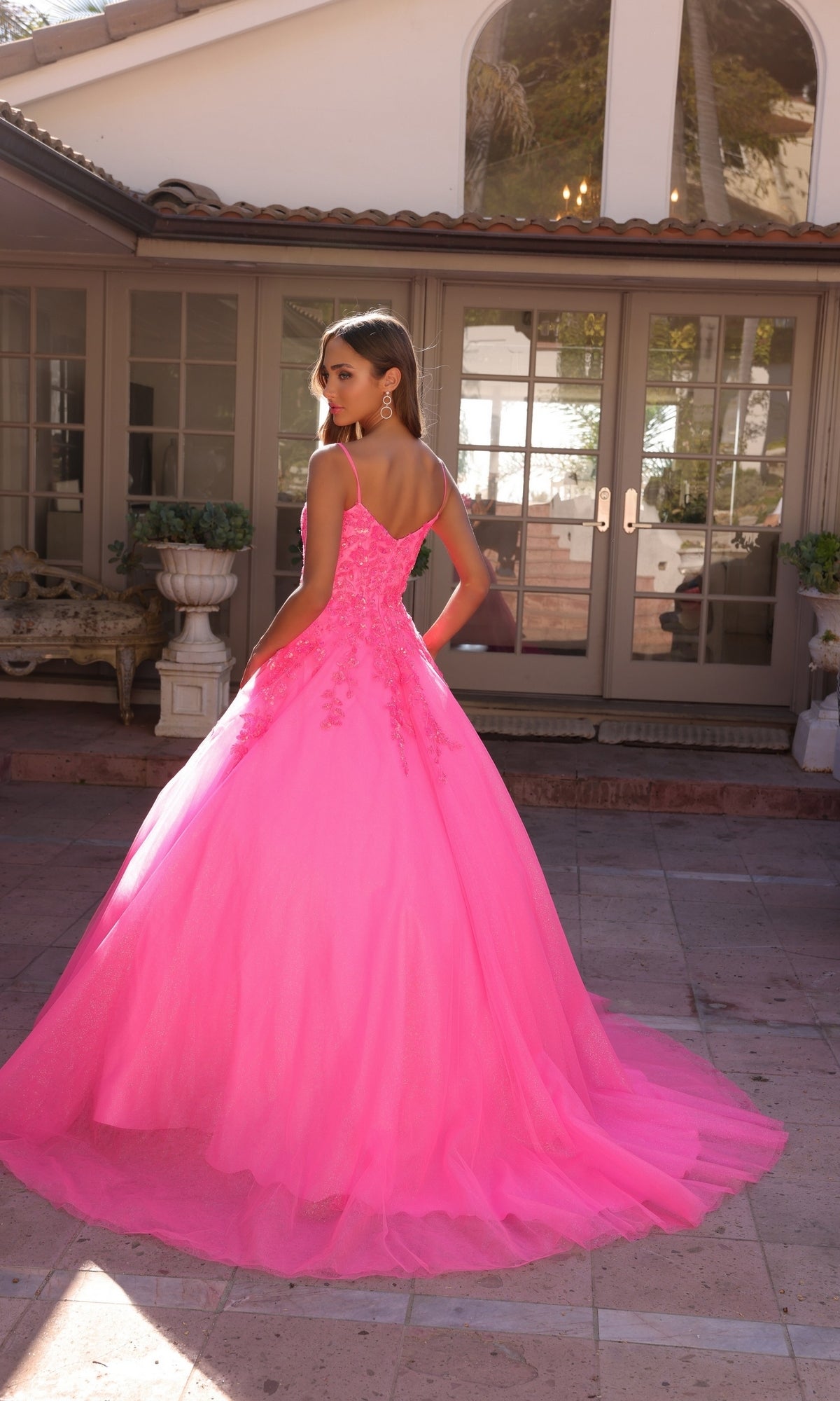 A woman in the Off-the-Shoulder Lace-Up Long Prom Ball Gown H1352, featuring vibrant pink embroidered lace, stands on a stone patio facing a building with large glass doors, her back to the camera.